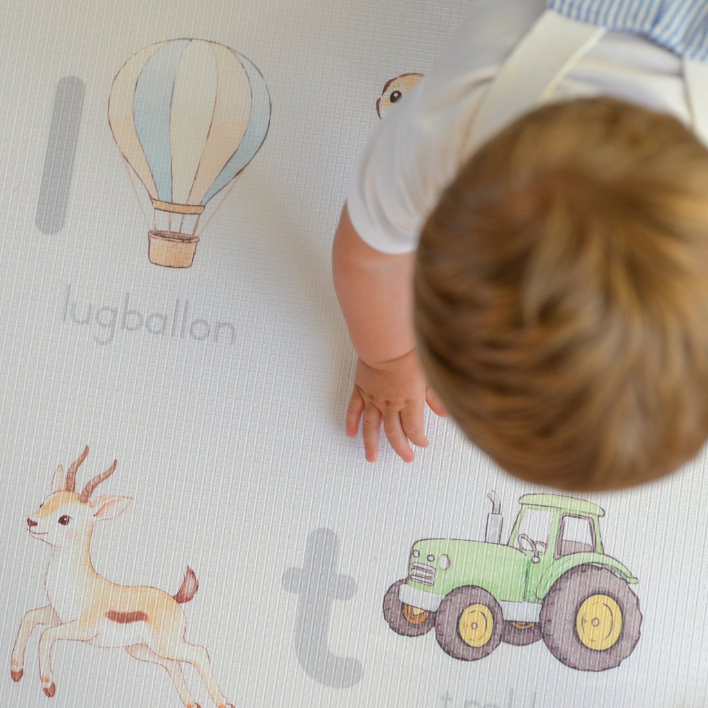 Child playing on an Afrikaans alphabet play mat featuring illustrations of a hot air balloon, Springbok, and tractor.