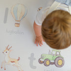Child playing on an Afrikaans alphabet play mat featuring illustrations of a hot air balloon, Springbok, and tractor.