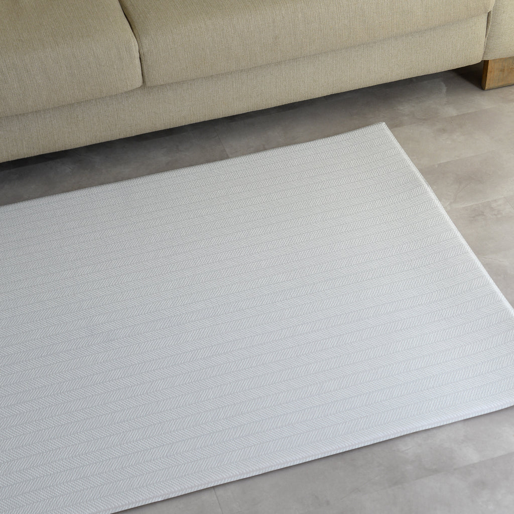 A modern play mat with a sage herringbone pattern on a gray floor in a room with a beige sofa.