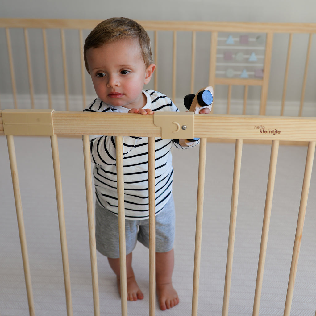 Child standing in a wooden baby playpen with built-in toys in a room.