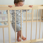 Child standing on a play mat inside a wooden playpen with a neutral background