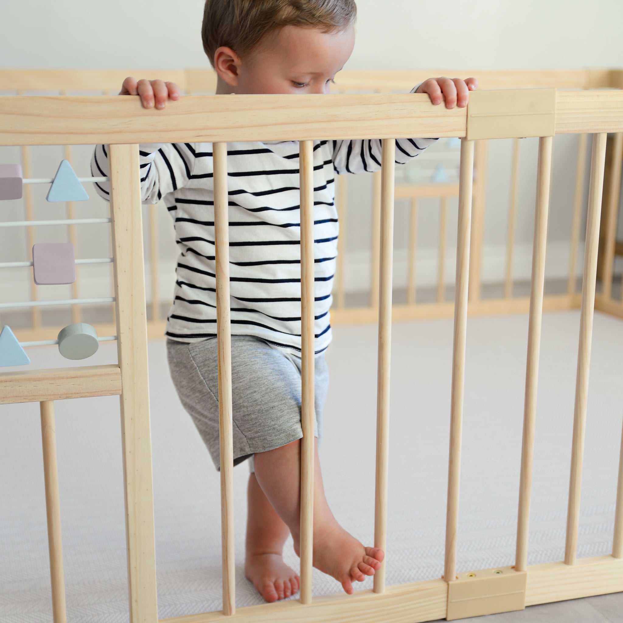 Child standing on a play mat inside a wooden playpen with a neutral background