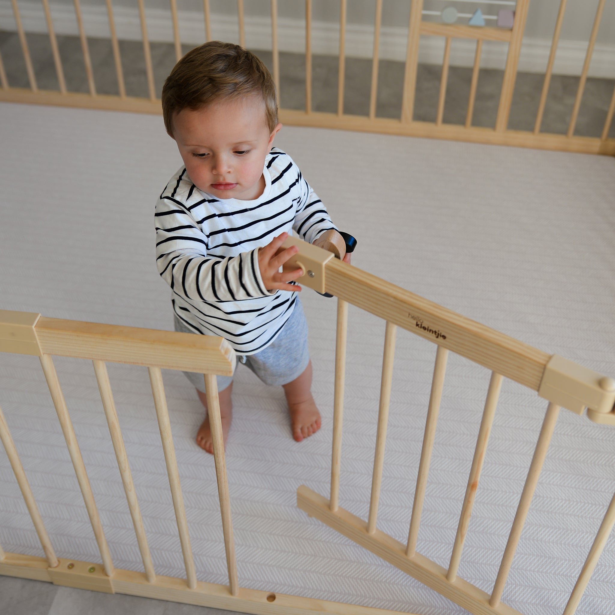 Child standing on a play mat in a wooden baby playpen in a room